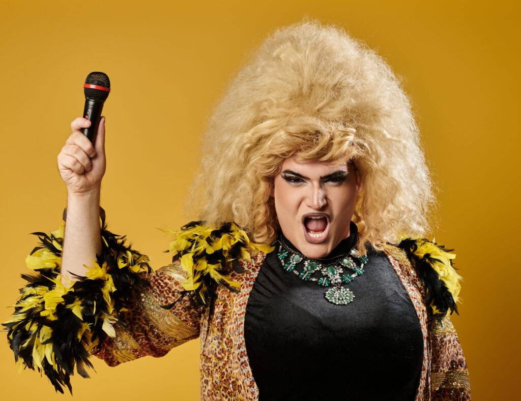 Libera la Tua Espressione 2 Una drag queen vestita in modo appariscente con una grande parrucca bionda, un'espressione accattivante e un microfono in mano